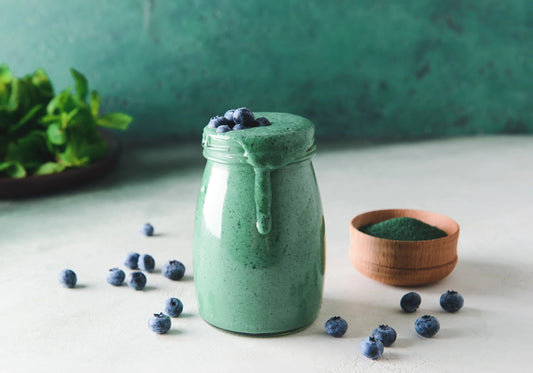 Green spirulina smoothie with blueberries in a glass jar on white table with spirulina powder in wooden bowl