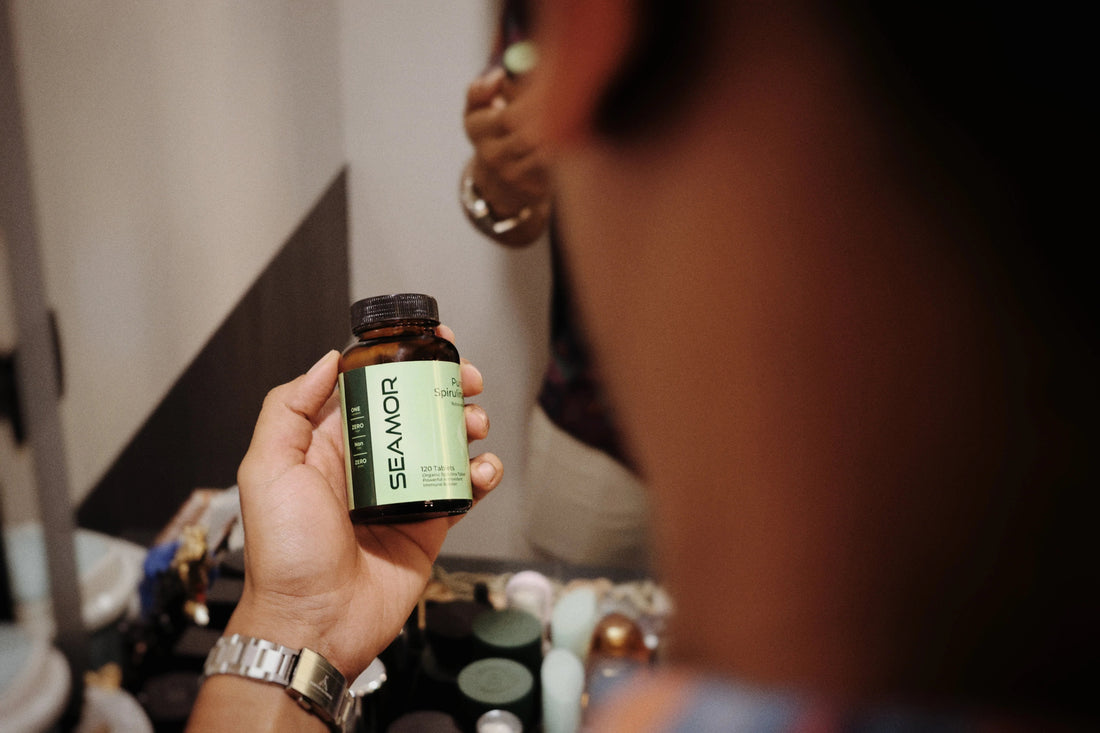 Hand holding a bottle of SEAMOR Pure Spirulina nutritional supplement with skincare products in the background