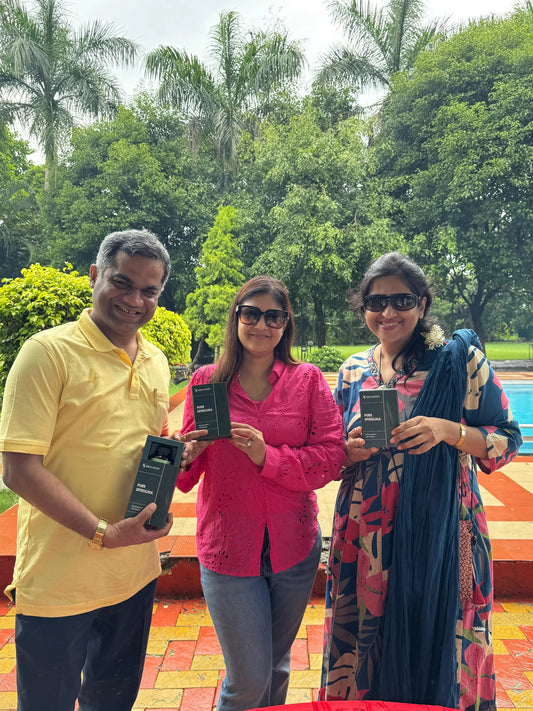 Three smiling adults outdoors holding Slimol Pure Spirulina supplement boxes near pool with lush greenery